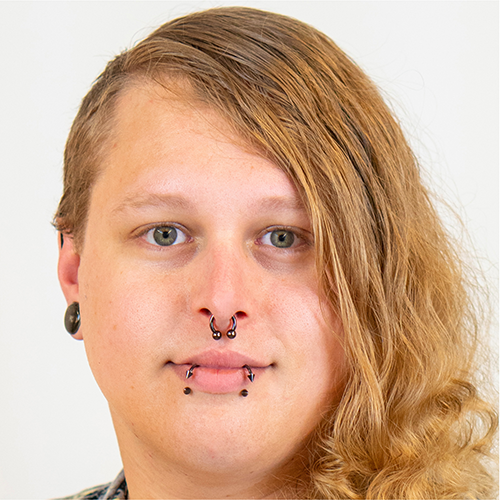 Portrait of Jacq Brasseur, a non-binary white person with long blonde hair and dark rimmed glasses. They are looking out of frame, to the left, with a small smile on their lips.